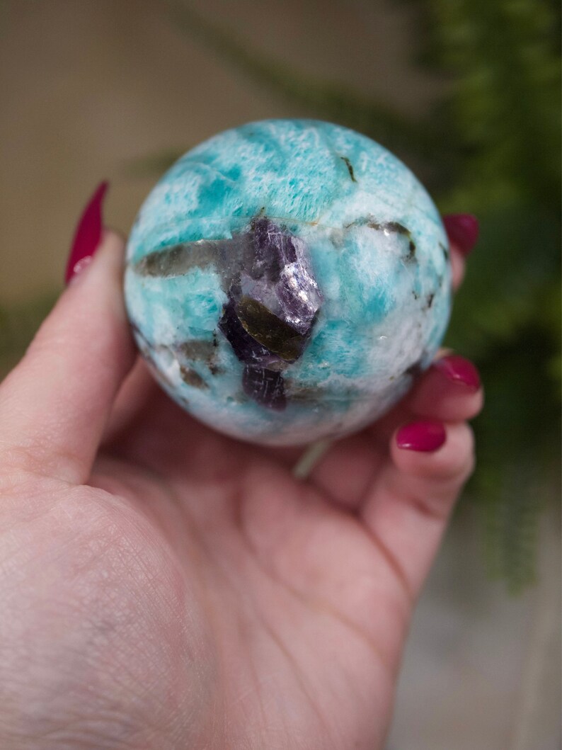 May include: A blue and green amazonite sphere with a purple amethyst inclusion. The sphere is smooth and polished.