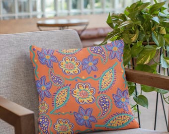 Può includere: Un cuscino decorativo con un motivo floreale colorato su sfondo arancione. Il cuscino è appoggiato su una sedia marrone con un cuscino in tessuto grigio.