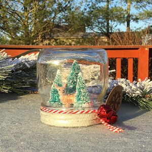 May include: A glass jar snow globe with a white base and red and white twine. Inside the globe is a miniature winter scene with two green trees and a brown deer. The snow globe is sitting on a sparkly white surface.