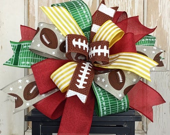 May include: A black lantern with a red, yellow, and green football-themed bow on top. The lantern has a yellow candle inside.