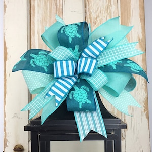 May include: A teal, blue, and white bow with a sea turtle print is tied around the top of a black lantern. The lantern has a yellow candle inside.