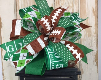 May include: A green, brown, and white football-themed bow sits atop a black lantern. The bow is made of ribbon with footballs, a football field, and a plaid pattern. The lantern has a candle inside and a copper leaf decoration.