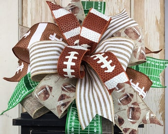 May include: A brown, white, and green football-themed bow with a burlap ribbon and a green and white ribbon with football field markings. The bow is on top of a black lantern with a yellow candle inside.