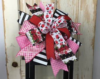 Könnte beinhalten: Eine rosa, rote, schwarze und weiße Schleife mit Blumen- und Tupfenmustern. Die Schleife ist um eine schwarze Laterne gebunden.