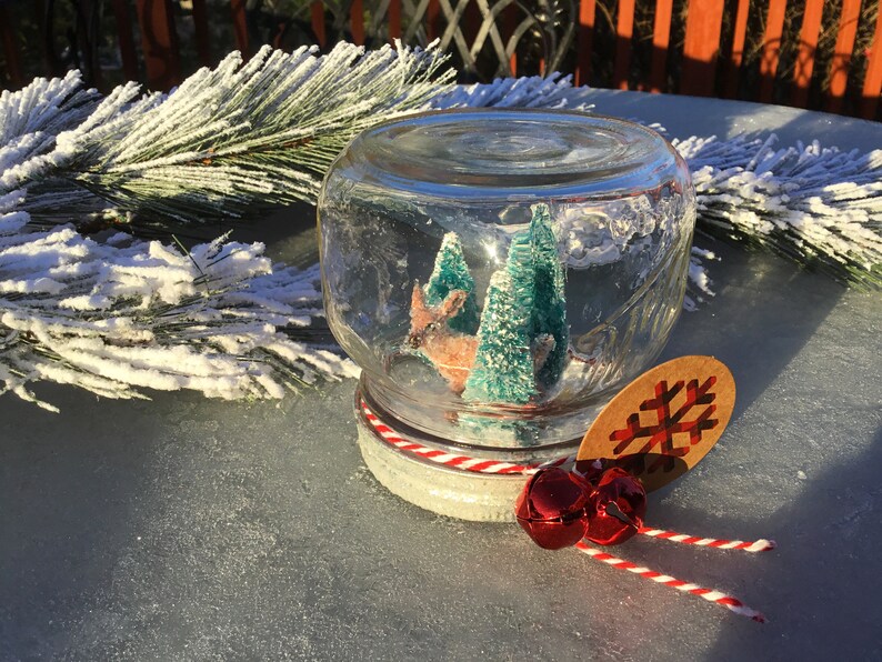 May include: A glass snow globe with a red and white striped ribbon around the lid. Inside the globe, there is a small deer figurine and two green pine trees. A brown paper tag with a red snowflake is attached to the ribbon.