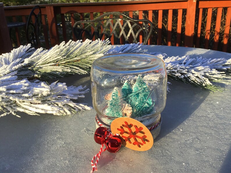 May include: A glass jar snow globe with a brown lid and a snowflake tag. Inside the globe are two small green glitter trees. The snow globe is sitting on a table with a white snowy pine branch.
