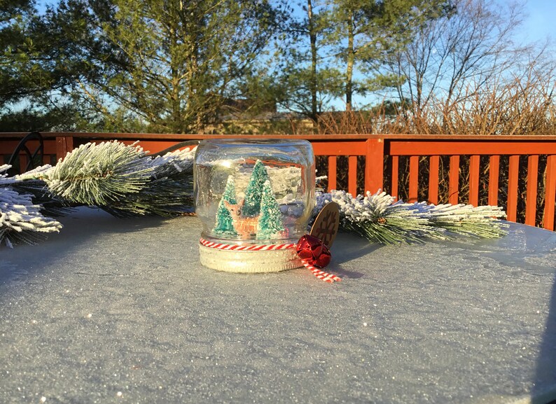 May include: A glass jar snow globe with a white base and red and white twine. Inside the globe is a miniature winter scene with two green trees and a brown deer. The snow globe is sitting on a sparkly white surface.