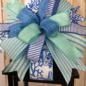 K&ouml;nnte beinhalten: Eine blaue und gr&uuml;ne gestreifte Schleife mit einem wei&szlig;en Band mit einem Seepferdchen- und Korallenmuster. Die Schleife ist um eine schwarze Laterne gebunden.