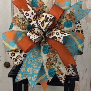 May include: A decorative fall bow featuring a mix of patterned ribbons. The bow includes ribbons with leopard print, teal and gold glitter, orange plaid, and pumpkin designs. The bow is a mix of orange, teal, brown, and cream colors.
