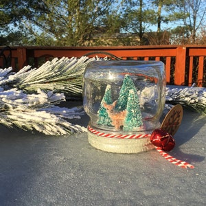 May include: A glass snow globe with a white and red striped ribbon, a red bell, and a snowflake tag. Inside the globe are two small green trees and a white deer figurine.