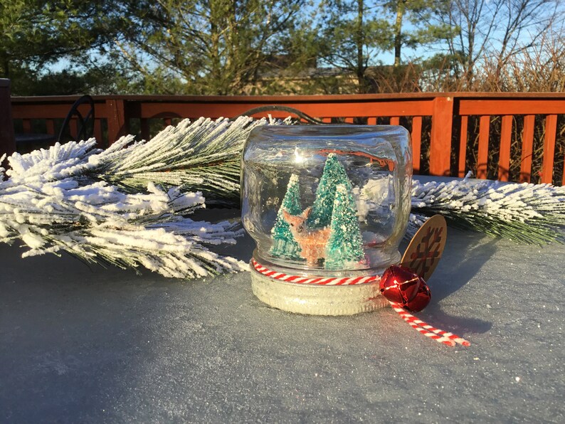 May include: A glass snow globe with a white and red striped ribbon, a red bell, and a snowflake tag. Inside the globe are two small green trees and a white deer figurine.