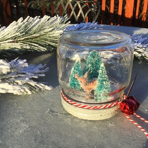 May include: A glass jar snow globe with a white base and red and white striped ribbon. Inside the globe are two small green trees and a pink deer figurine.