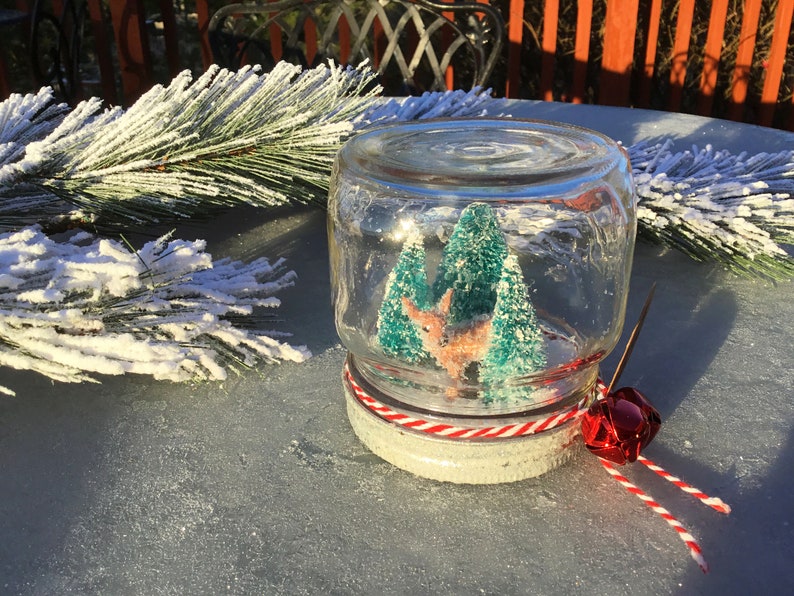 May include: A glass jar snow globe with a white base and red and white striped ribbon. Inside the globe are two small green trees and a pink deer figurine.