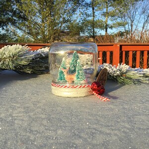 May include: A glass snow globe with a red and white striped ribbon and a red bell. Inside the globe is a miniature deer and two green trees. The snow globe is sitting on a white surface.