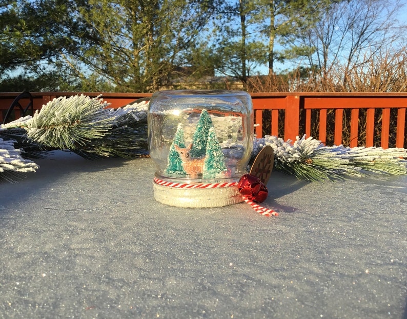 May include: A glass snow globe with a red and white striped ribbon and a red bell. Inside the globe is a miniature deer and two green trees. The snow globe is sitting on a white surface.
