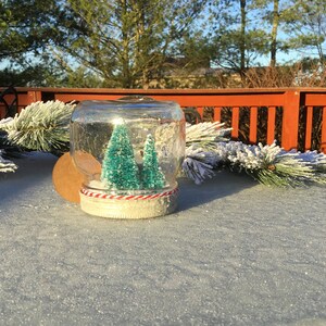 May include: A glass jar snow globe with two small green artificial Christmas trees inside. The jar is decorated with a red and white striped ribbon around the base.