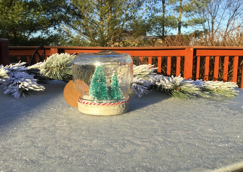 May include: A glass jar snow globe with two small green artificial Christmas trees inside. The jar is decorated with a red and white striped ribbon around the base.