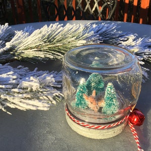May include: A glass jar snow globe with a red and white striped lid and a red bell attached to the lid. Inside the snow globe are two small green trees and a small brown deer figurine.