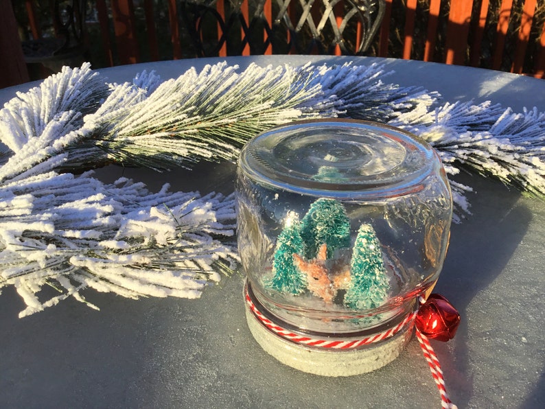 May include: A glass jar snow globe with a red and white striped lid and a red bell attached to the lid. Inside the snow globe are two small green trees and a small brown deer figurine.
