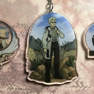 May include: Three clear acrylic keychains featuring illustrated characters and a robot. The left keychain shows two figures and a sword. The center keychain depicts a person in a white coat. The right keychain shows a robot with the text happy beeping.