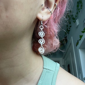 May include: Silver spiral dangle earrings with multiple loops. The earrings are hanging from a person's ear.