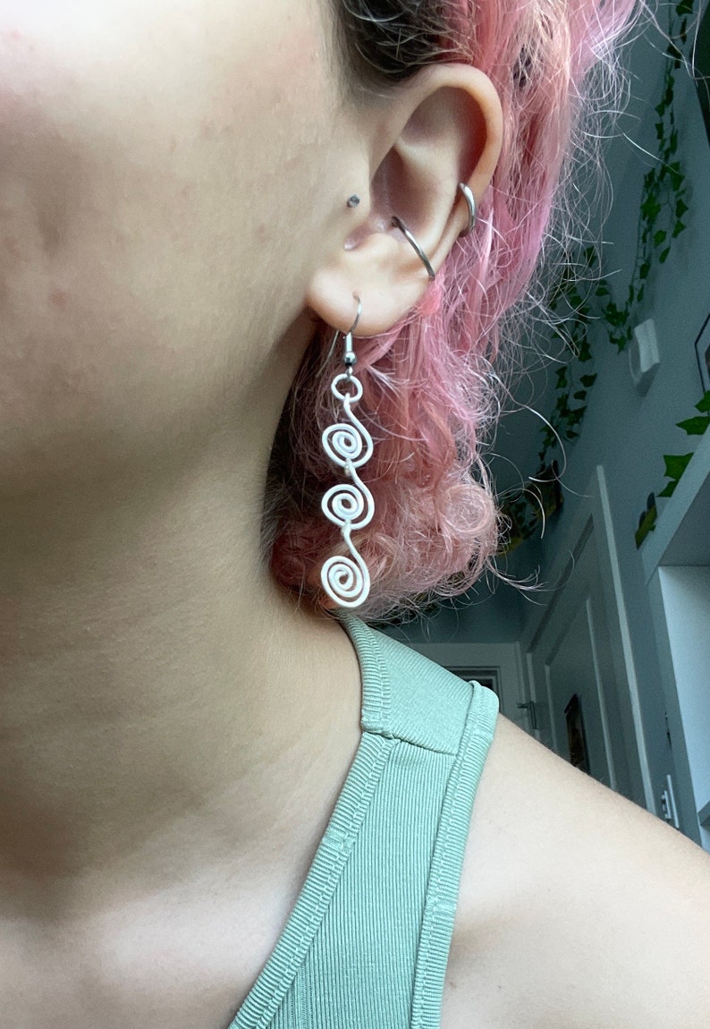 May include: Silver spiral dangle earrings with multiple loops. The earrings are hanging from a person's ear.
