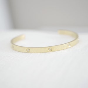 Könnte beinhalten: Ein goldenes Armreif-Armband mit kleinen Diamanten, die in die Band eingefasst sind.