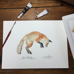 May include: Watercolor painting of a red fox jumping in the air, with a white background. The painting is on a white mat and is surrounded by a wooden frame. There are paint tubes and a palette with paint on the table.