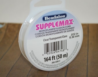 May include: A spool of clear Beadalon Supplemax monofilament illusion cord. The spool is labelled with the text "Supplemax", "Monofilament Illusion Cord", "Cordon Transparent Monofilament", "Cordon Monofilamento de Ilusion", "Clear/Transparent/Claro", "0.012 in", "0.30 mm", "164 ft (50 m)", and "JNO.30W-F".