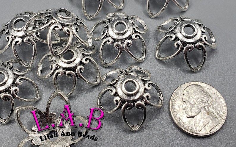 K&ouml;nnte beinhalten: Eine Sammlung silberfarbener Metallperlenkappen mit floralem Design. Die Kappen haben ein zentrales kreisf&ouml;rmiges Element, das von sechs bl&uuml;tenblattf&ouml;rmigen Schlaufen umgeben ist. Eine US-Nickel-M&uuml;nze dient als Gr&ouml;&szlig;enreferenz. Der Text "L.A.B Lilah Ann Beads" ist ebenfalls enthalten.
