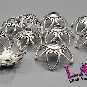 K&ouml;nnte beinhalten: Silberfarbene Metallperlenkappen mit floralem Design. Die Kappen haben ein zentrales Loch und sechs gebogene Bl&uuml;tenbl&auml;tter mit dekorativen Wirbeln. Das Bild enth&auml;lt den Text "L.A.B Lilah Ann Beads".