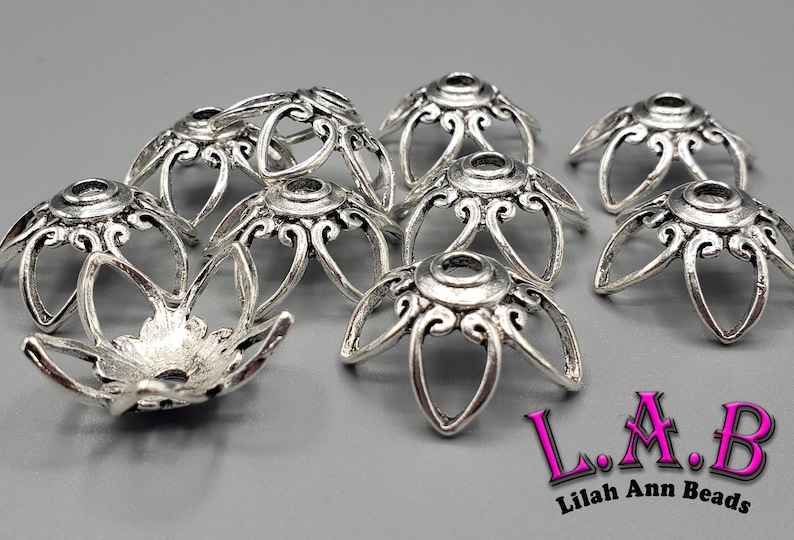 K&ouml;nnte beinhalten: Silberfarbene Metallperlenkappen mit floralem Design. Die Kappen haben ein zentrales Loch und sechs gebogene Bl&uuml;tenbl&auml;tter mit dekorativen Wirbeln. Das Bild enth&auml;lt den Text "L.A.B Lilah Ann Beads".