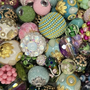 May include: A collection of colorful beads in various shapes and sizes. The beads are in shades of pink, blue, green, and gold, with intricate designs and textures. Some beads have a textured surface, while others are smooth and shiny. The beads are likely for jewelry making.