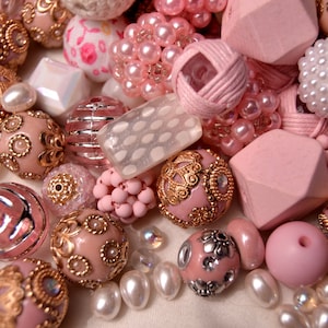 May include: A collection of pink and white beads with gold accents. The beads are made from a variety of materials, including wood, glass, and metal. Some of the beads feature intricate designs, while others are plain. The beads are ideal for making jewellery or other crafts.