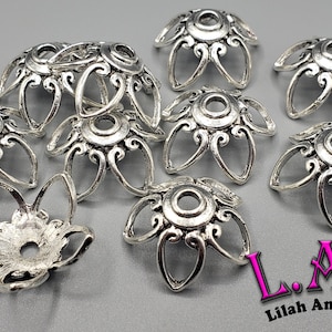 K&ouml;nnte beinhalten: Silberfarbene Metallperlenkappen mit floralem Design. Die Kappen haben ein zentrales Loch und sind von stilisierten Bl&uuml;tenbl&auml;ttern umgeben. Das Bild enth&auml;lt den Text "L.A.B Lilah Ann Beads".