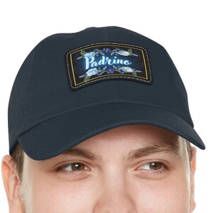 May include: A navy blue baseball cap with a rectangular patch. The patch features the word "Padrino" in white script, surrounded by blue floral designs on a gold background. The cap is made of a soft material.
