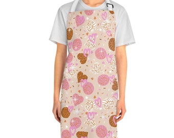 May include: Apron with a beige background featuring a pattern of pink and brown concha bread, pink hearts, and sprinkles. The apron has a white neck strap and a black adjustable buckle.
