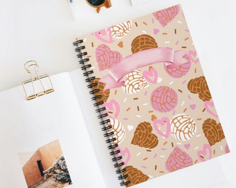 May include: A spiral-bound notebook with a beige cover featuring a pattern of pink and brown concha bread shapes, hearts, and sprinkles. A pink ribbon is across the top. The notebook is open on a white surface.