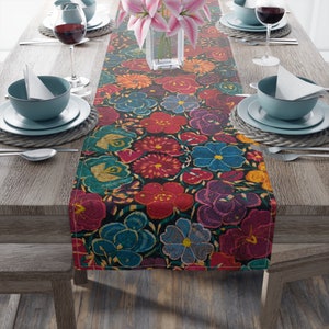 May include: A colourful floral table runner featuring a pattern of red, blue, purple, and yellow flowers against a dark background. The runner is on a wooden table set for a meal, with plates, cutlery, and wine glasses.