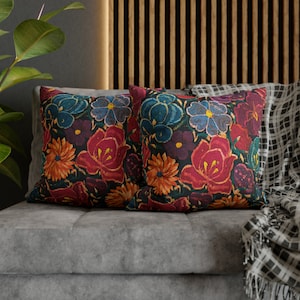 May include: Two square cushions featuring a dark floral design with red, orange, blue, and purple flowers. The cushions are placed on a grey sofa, with a patterned blanket visible. The cushions would measure approximately 40cm x 40cm.