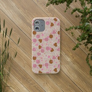 May include: A phone case with a beige background featuring a pattern of pink and brown conchas, hearts, and sprinkles. The case has a camera cutout and is on a wooden surface.