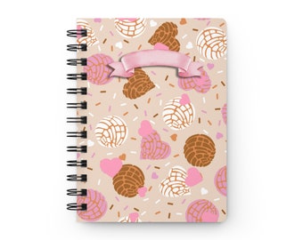 May include: Spiral-bound notebook featuring a repeating pattern of concha bread, hearts, and sprinkles in shades of pink, brown, and white on a beige background. A pink ribbon banner is across the top. The notebook has a black spiral binding.