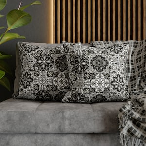 May include: Two square throw pillows with a black and white tile pattern featuring floral and geometric designs. The pillows are placed on a gray sofa, with a checkered blanket visible on the right side.