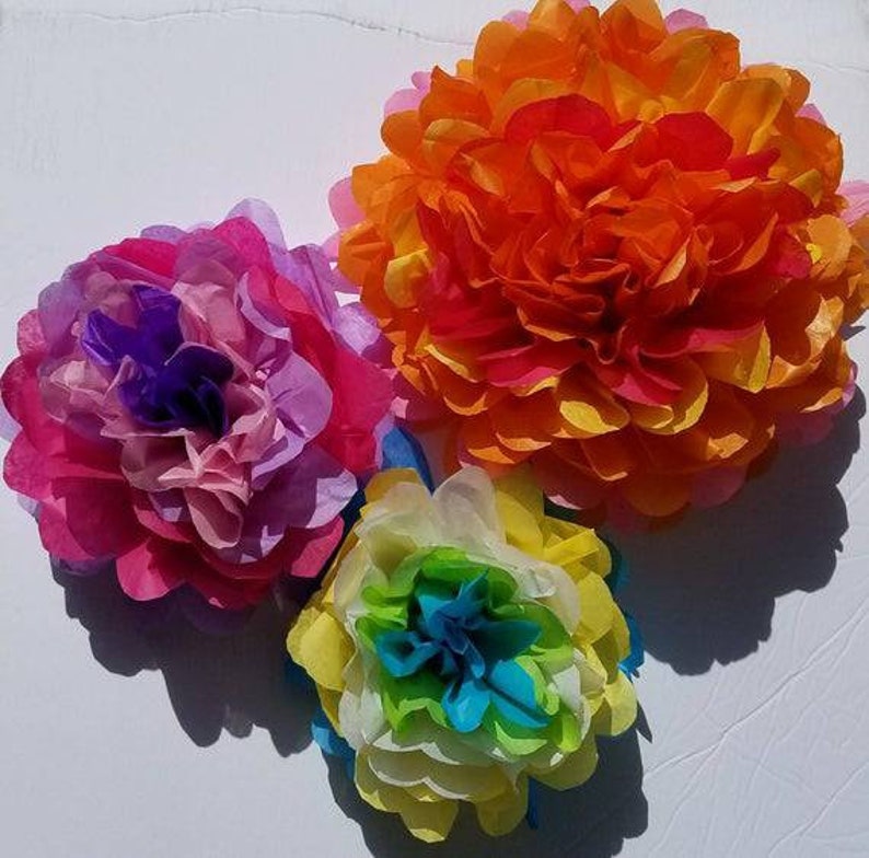 May include: Three colorful tissue paper flowers in shades of pink, orange, yellow, and blue. The flowers are arranged on a white surface, with shadows adding depth. The flowers have a layered petal design.