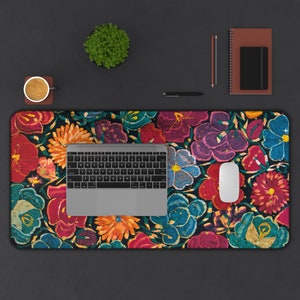 Pode incluir: Tapete de mesa com padrão floral com laptop, mouse e teclado. O tapete apresenta flores bordadas em vermelho, roxo, azul e laranja sobre um fundo escuro. Uma pequena planta, uma xícara de café, uma caneta e um caderno também estão presentes.