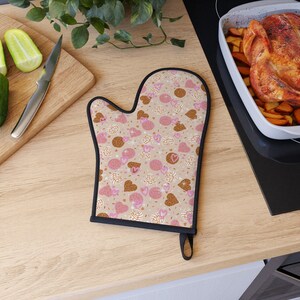 May include: Oven mitt in tan with a black trim and loop. It has a repeating pattern of pink and brown concha bread, hearts, and sprinkles. The mitt is designed for kitchen use.