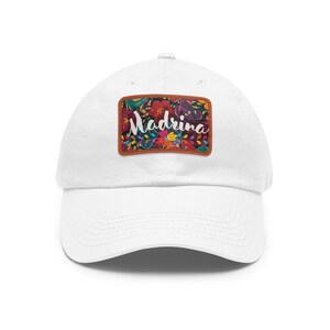 May include: White baseball cap with a colorful floral patch. The patch features the word "Madrina" in cursive, surrounded by vibrant flowers in red, purple, yellow, and blue. The cap is a classic six-panel design.