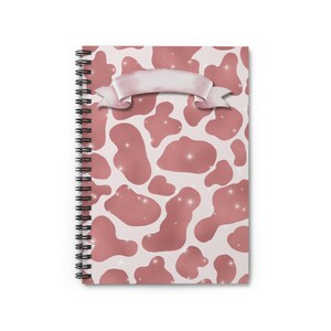 May include: A spiral-bound notebook with a pink cow print design. The cover features a light pink background with darker pink irregular shapes and a decorative banner. The design includes sparkling accents.