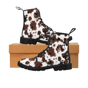 May include: Combat boots with a cow print design in brown and white, featuring black laces and soles. The boots have a pull tab at the back and are decorated with gold star accents.