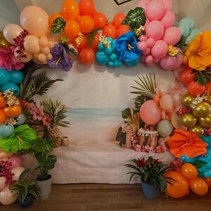 K&ouml;nnte beinhalten: Ein tropischer Ballonbogen mit Hintergrundbild einer Strandszene, verziert mit orangefarbenen, rosa und blauen Luftballons, gro&szlig;en Papierblumen, k&uuml;nstlichen Palmbl&auml;ttern und einer Ananas f&uuml;r eine lebendige, festliche Dekoration.